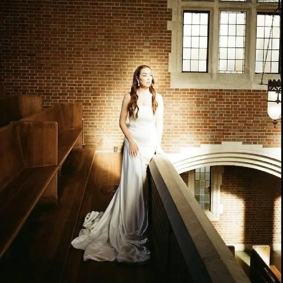 Woman in a white dress standing in a dimly lit room with large windows.