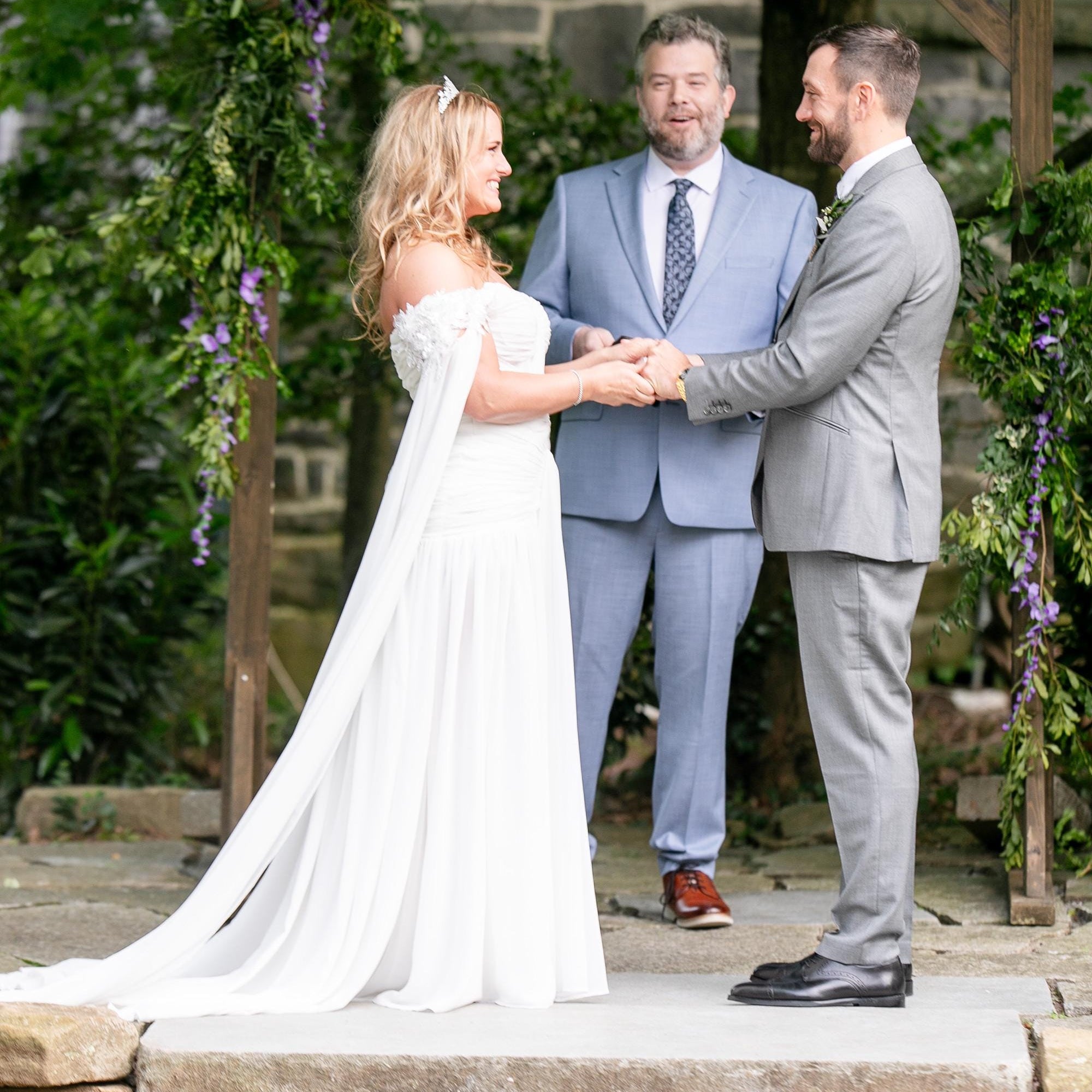 Wedding ceremony with a couple holding hands in front of an archway with greenery.