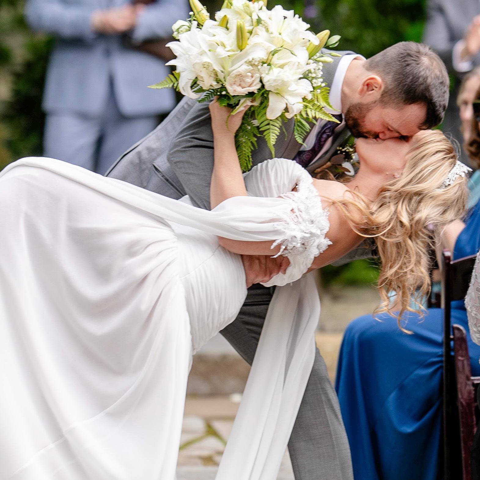 Man in a suit lifting a woman in a white dress, surrounded by people outdoors.