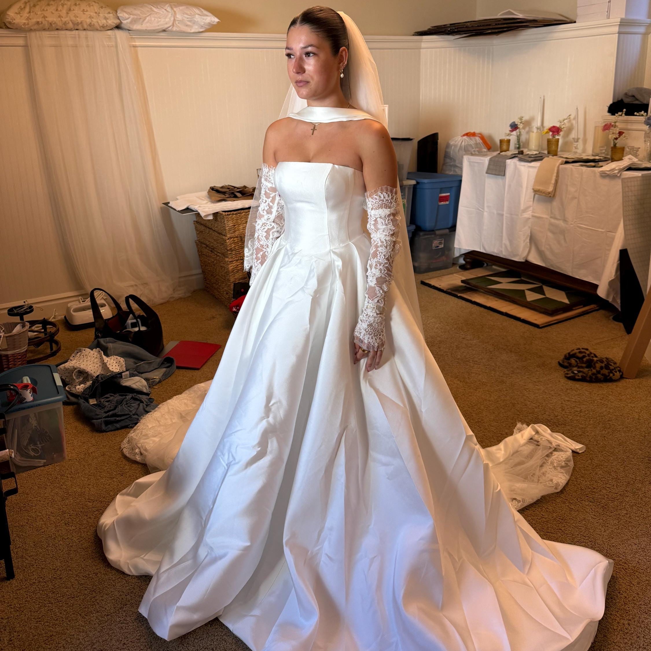 Woman in a white wedding dress standing in a room with various items scattered around.